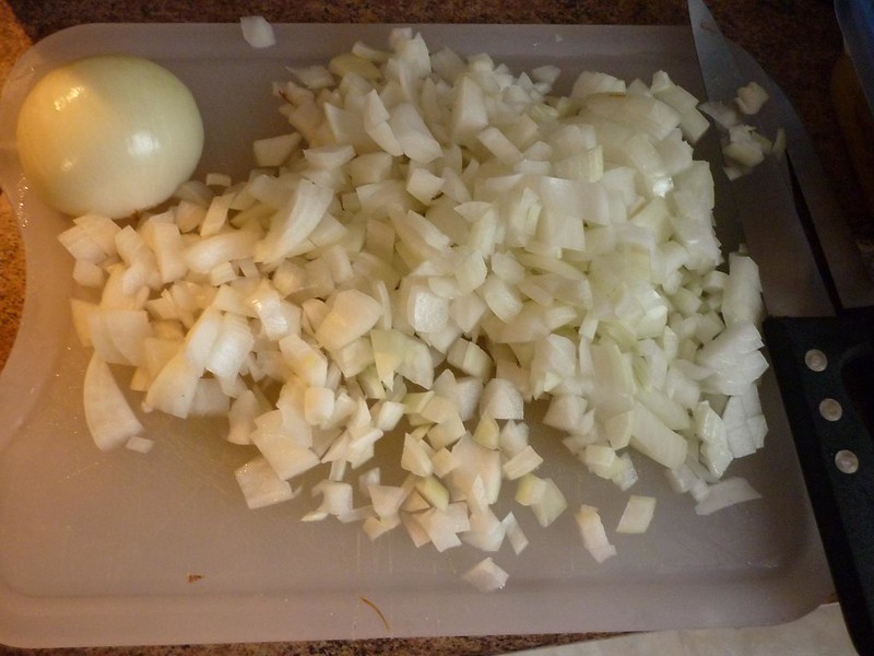 Cutting board with half an onion in the background and a pile of diced onions in the middle.